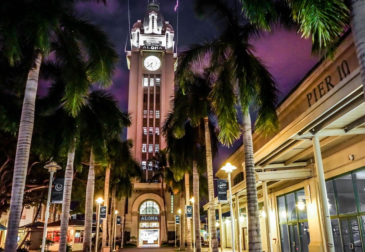 Night view of the Aloha Tower in Honolulu with palm trees and illuminated buildings.