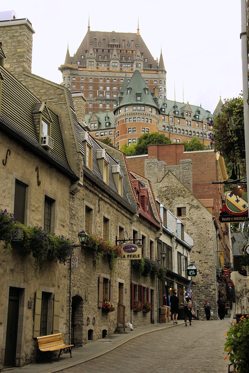 A narrow cobblestone street in a historic district with old stone buildings, flower boxes, and a large castle-like hotel in the background.