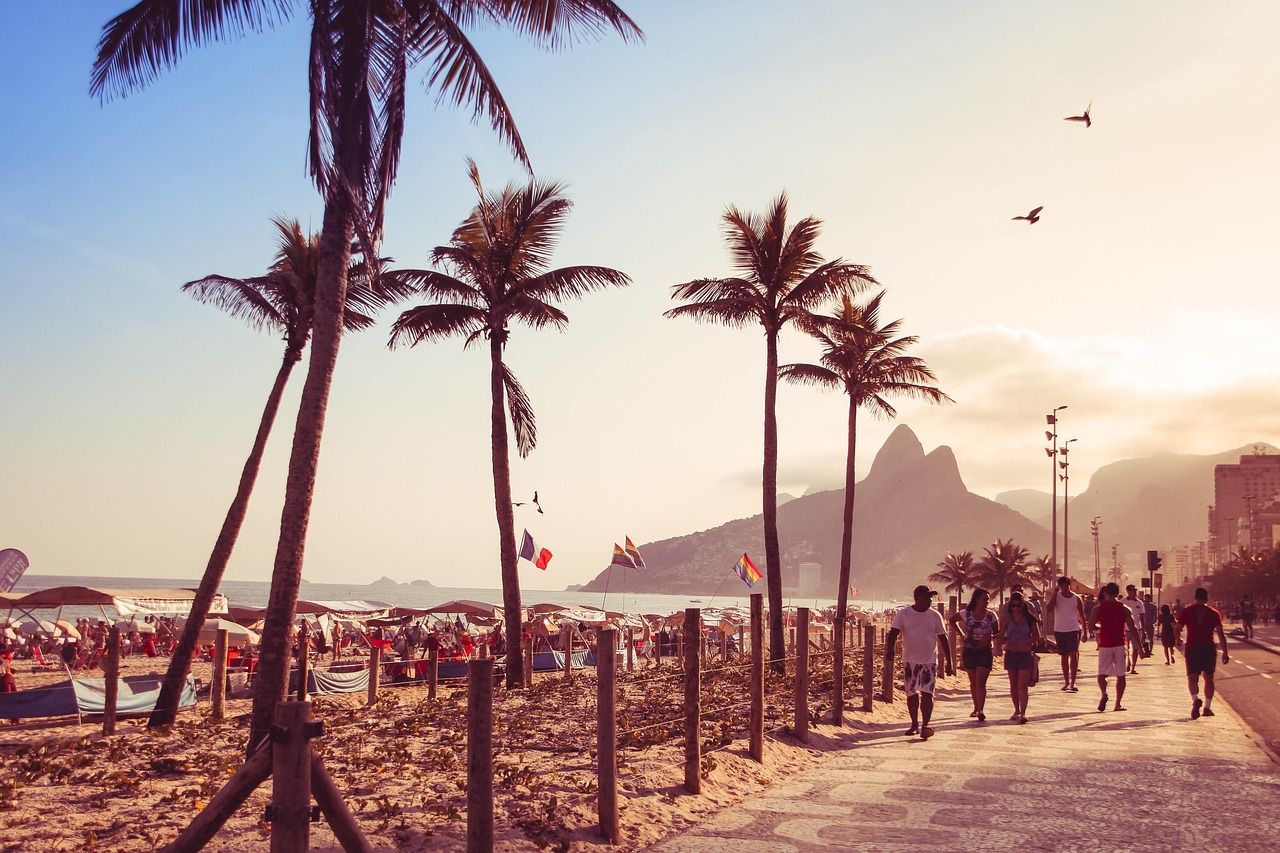 A scenic beach walkway with palm trees, people strolling, and a mountainous backdrop during sunset.