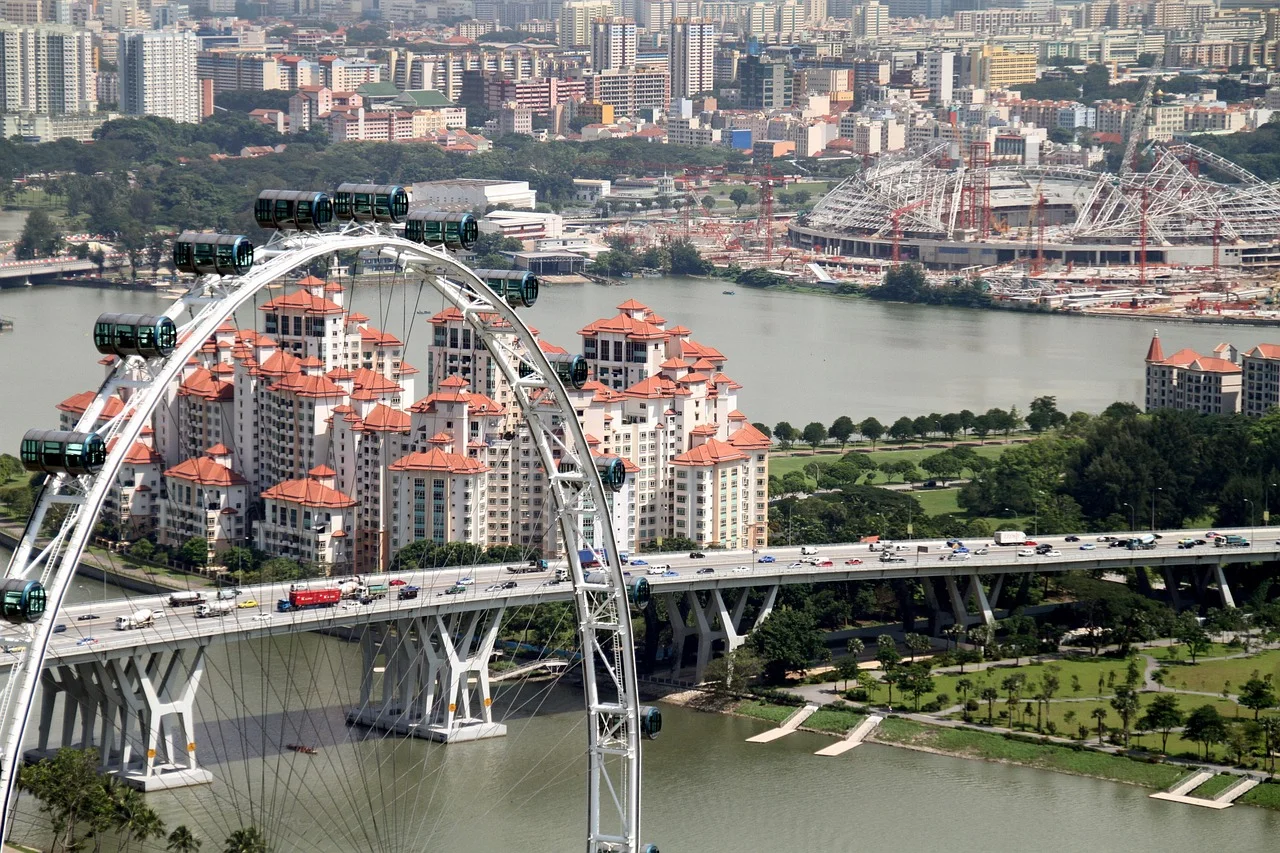 Aerial view of a city with a large Ferris wheel, residential buildings, a bridge, and a stadium near a body of water.