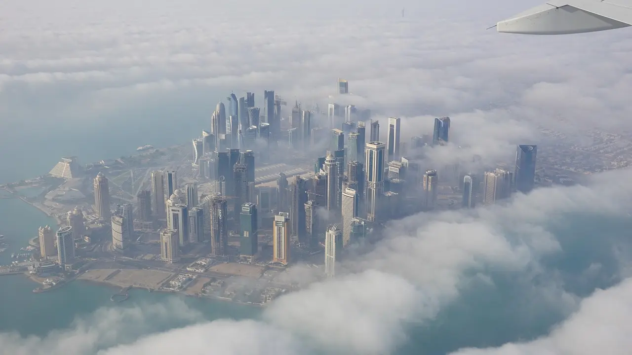 Aerial view of a modern city with tall skyscrapers and a waterfront, partially covered by clouds.