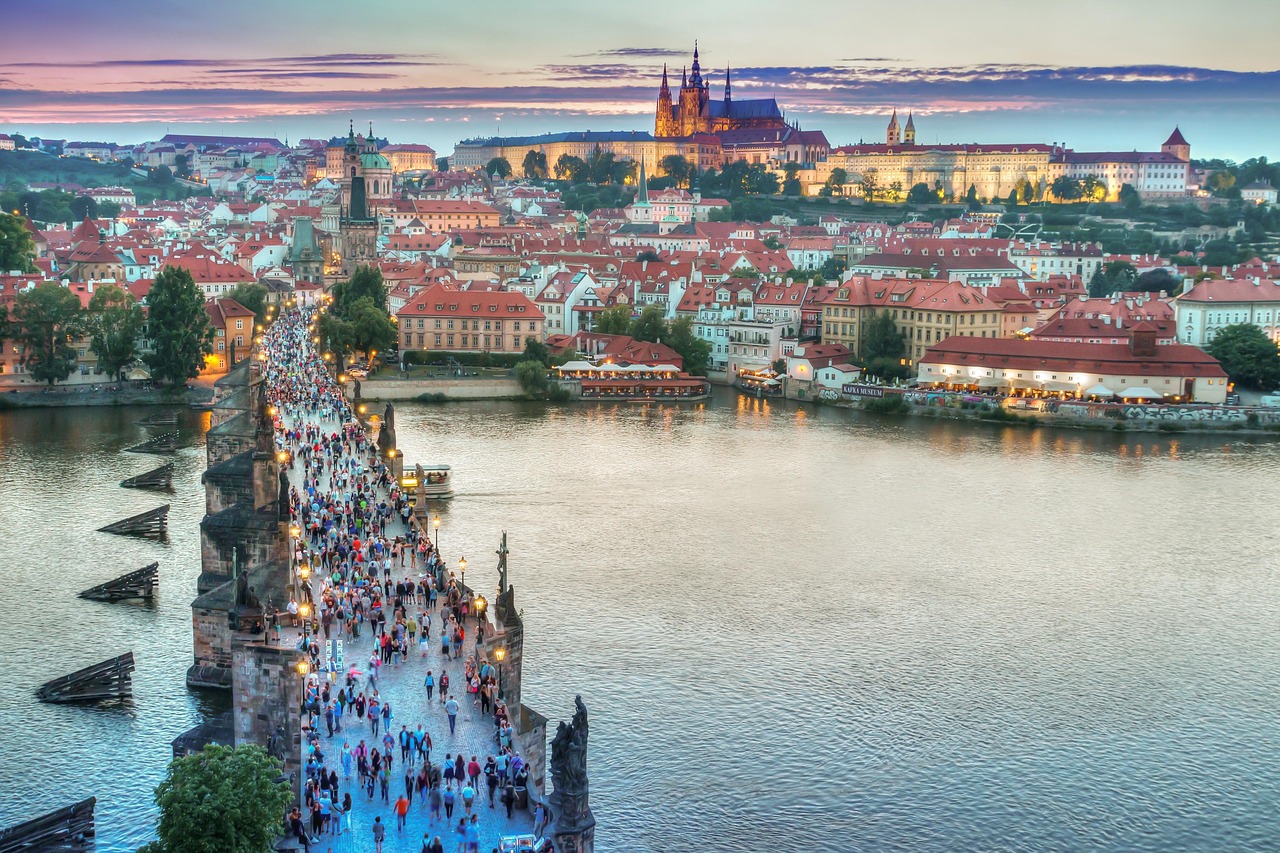 A bustling bridge over a river in Prague, Czechia, at dusk with people walking and historic buildings in the background.