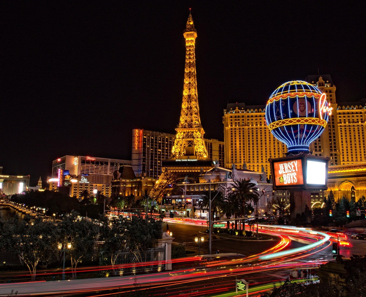 Night view of Las Vegas Strip with illuminated buildings, including a replica of the Eiffel Tower and a hot air balloon sign.