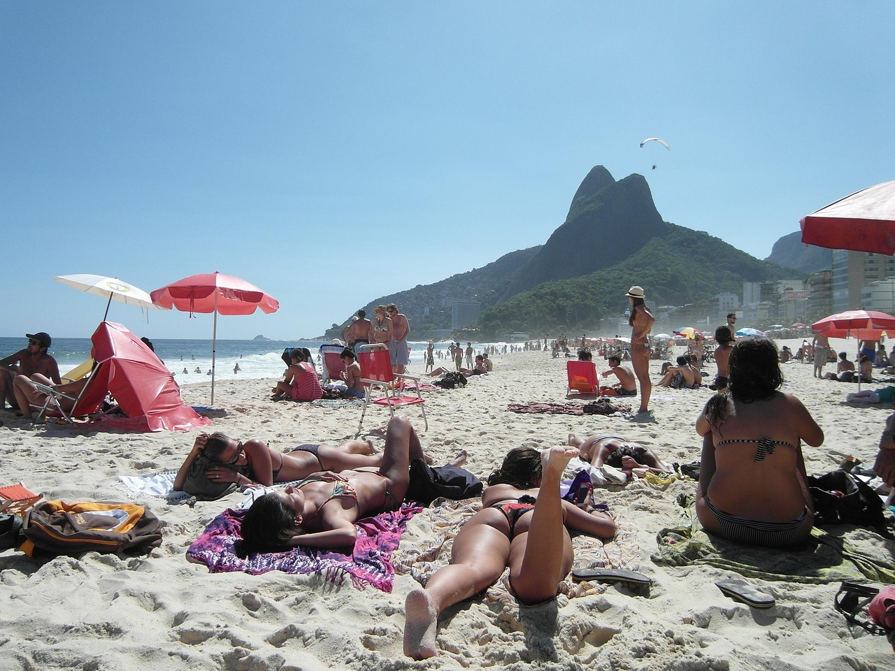 A crowded beach with people sunbathing and relaxing under umbrellas, with a prominent mountain in the background.