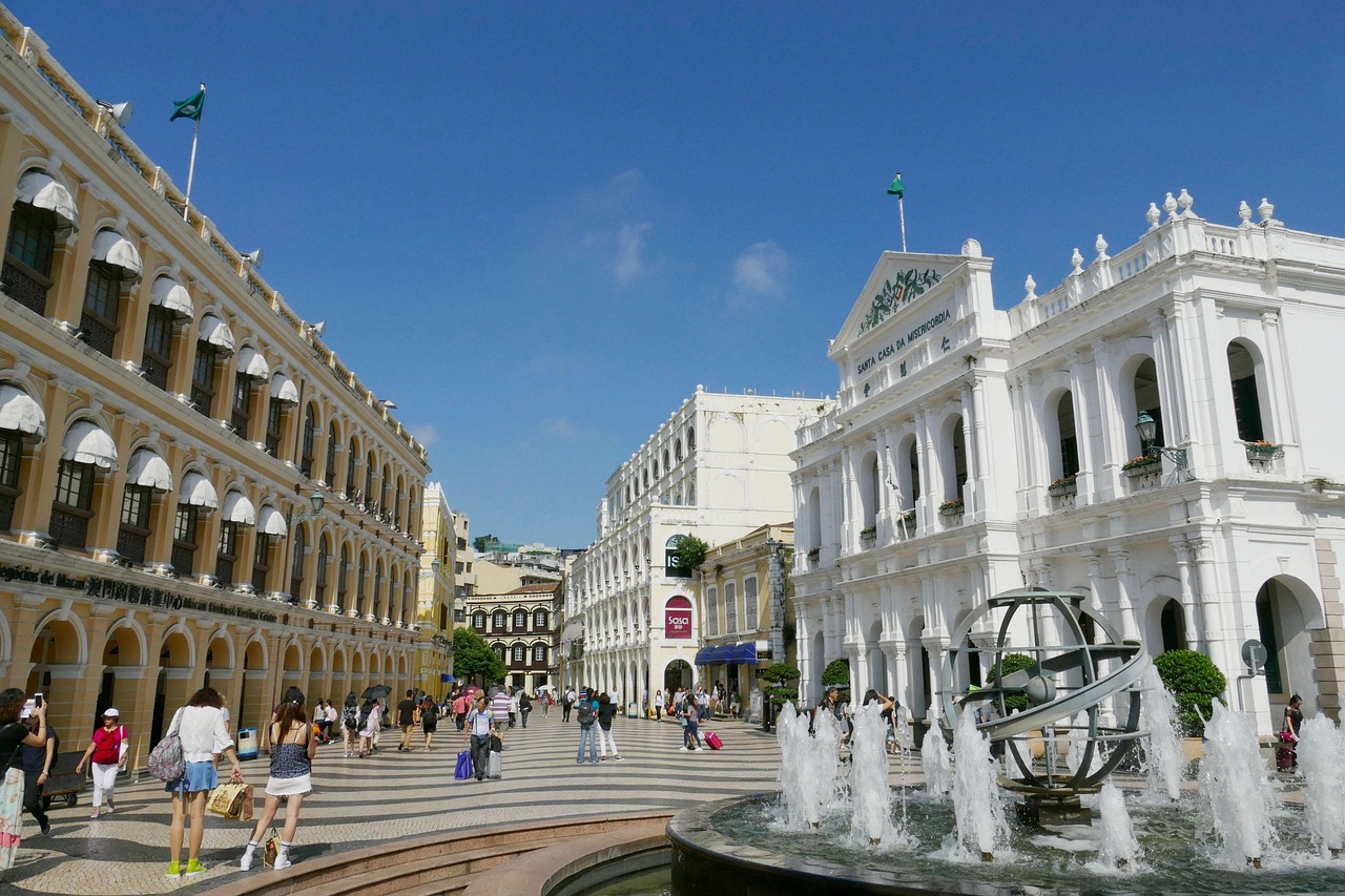 A bustling pedestrian street in Macau with historic buildings, a fountain, and people shopping and walking.