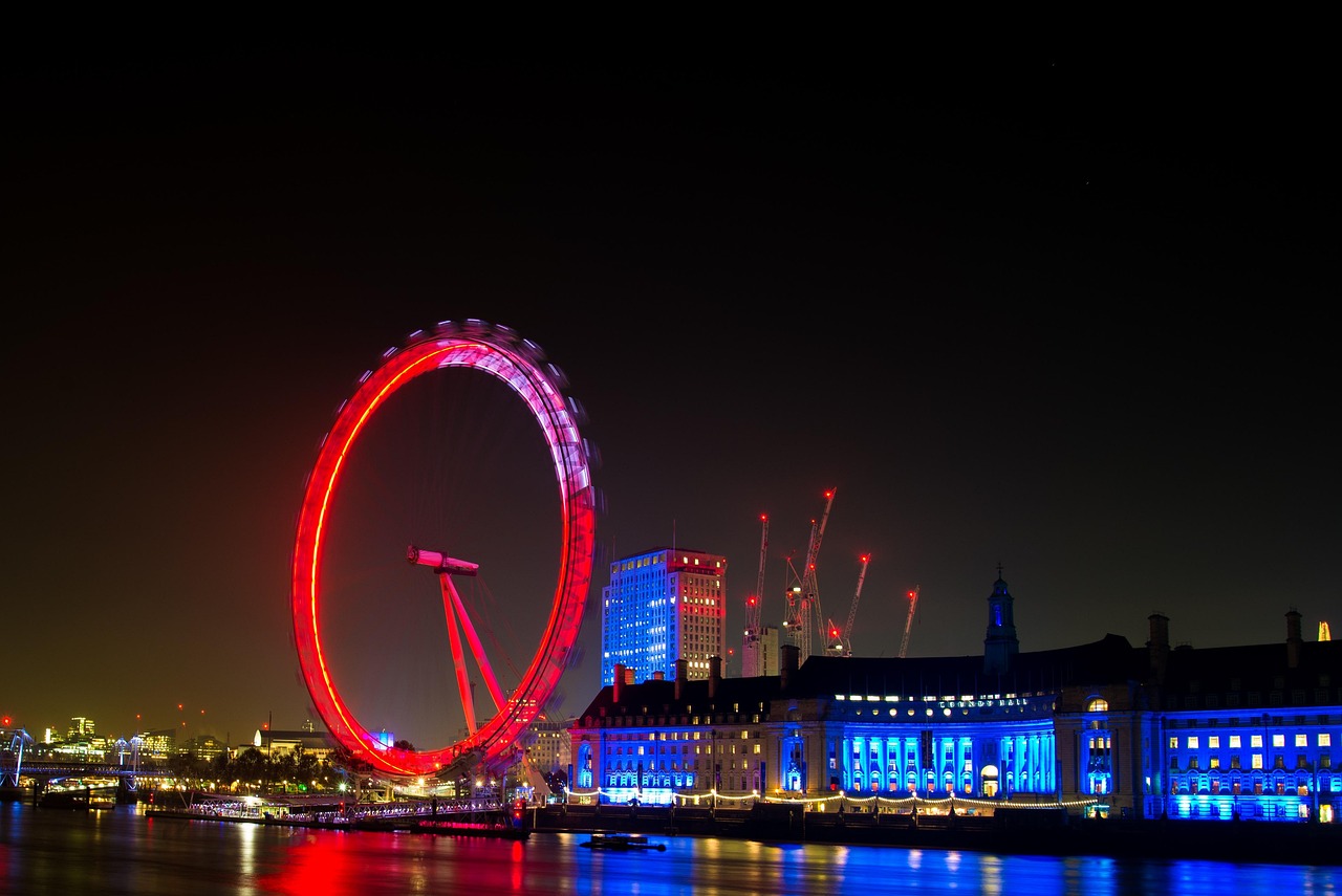 Night view of the London Eye and surrounding buildings illuminated with colorful lights, reflecting on the water.