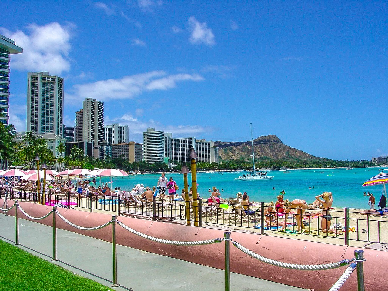 A bustling beach in Honolulu with tourists relaxing under umbrellas, high-rise buildings in the background, and a mountain in the distance.