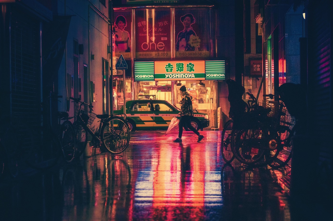 A nighttime street scene in an urban area with neon lights, a restaurant, bicycles, and a taxi.