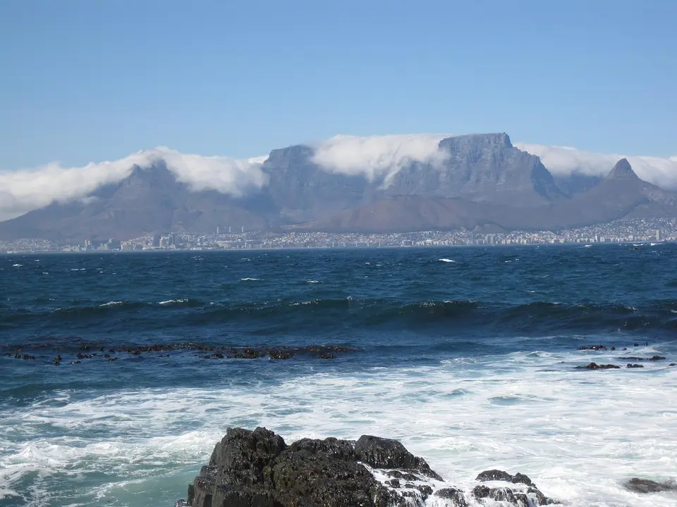 A view of Table Mountain with a cityscape in the background and waves crashing against rocks in the foreground.