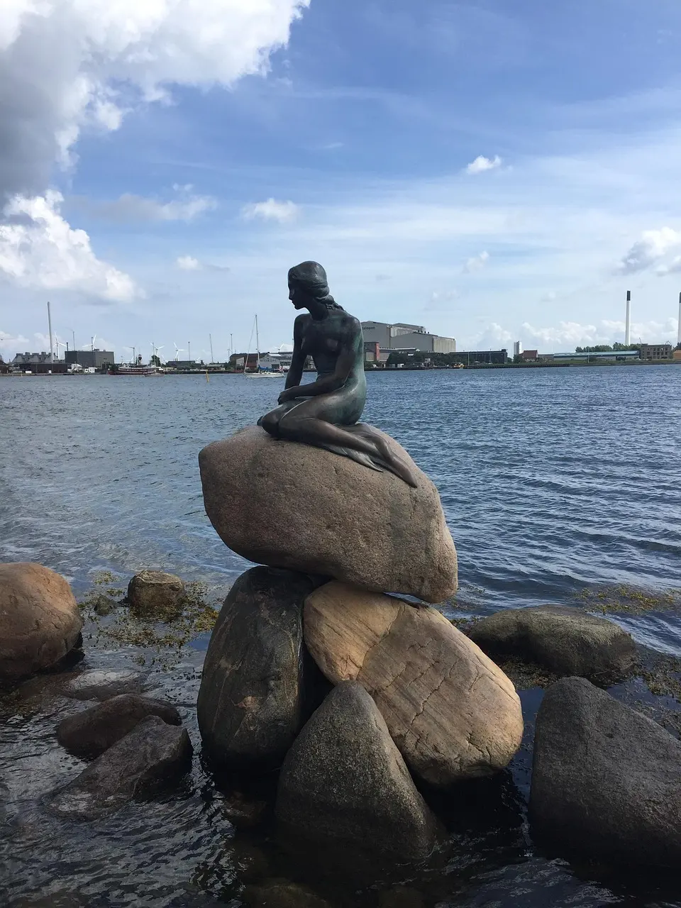 A statue of a mermaid sitting on a rock by the waterfront with an industrial harbor in the background.