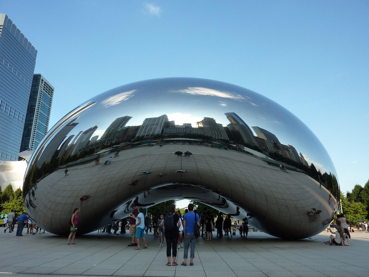 A large reflective bean-shaped sculpture in a city park with people around it and tall buildings reflected on its surface.