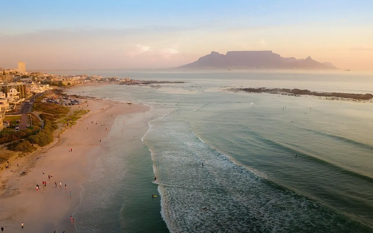 Aerial view of a beach with waves crashing onto the shore, people walking along the sand, and a cityscape with mountains in the background during sunset.