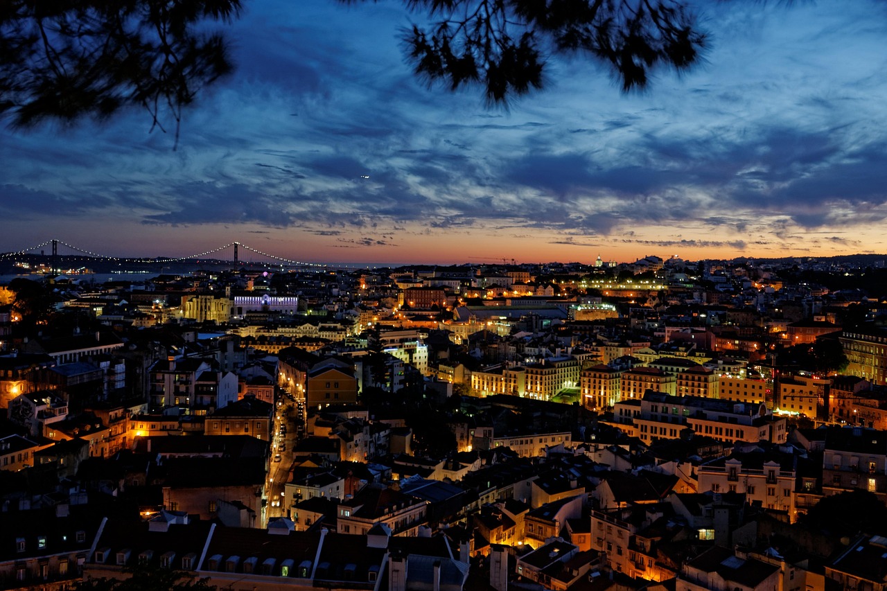 A nighttime cityscape view of Lisbon, Portugal, with illuminated buildings and a sunset sky.