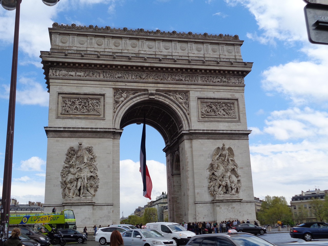 The Arc de Triomphe in Paris, France, with a French flag hanging from the top, cars and people around, and a clear blue sky.