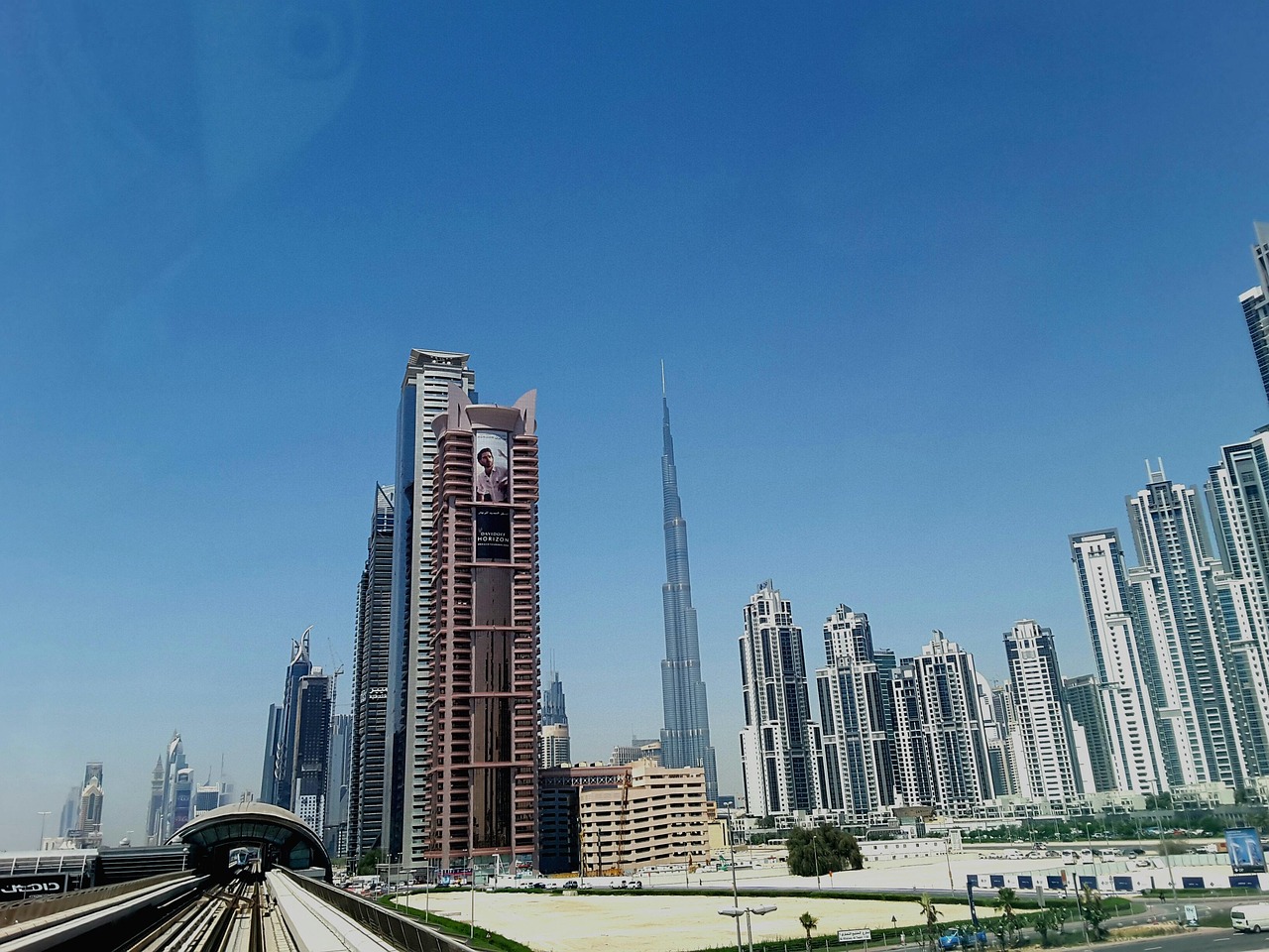 A cityscape featuring tall skyscrapers and a metro station in the foreground, with the Burj Khalifa prominently visible.