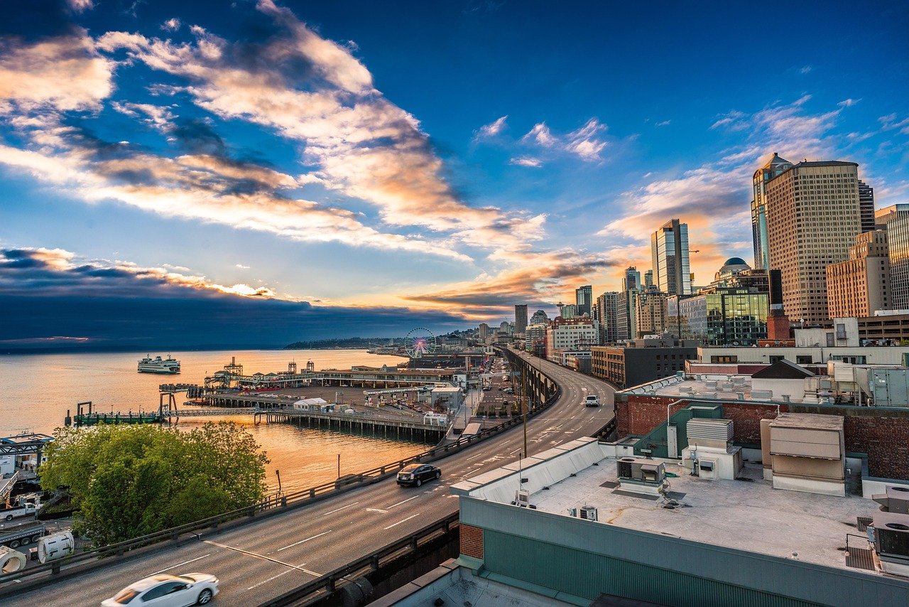 A cityscape at sunset with a highway running through it, leading to a waterfront with buildings and a Ferris wheel in the distance.