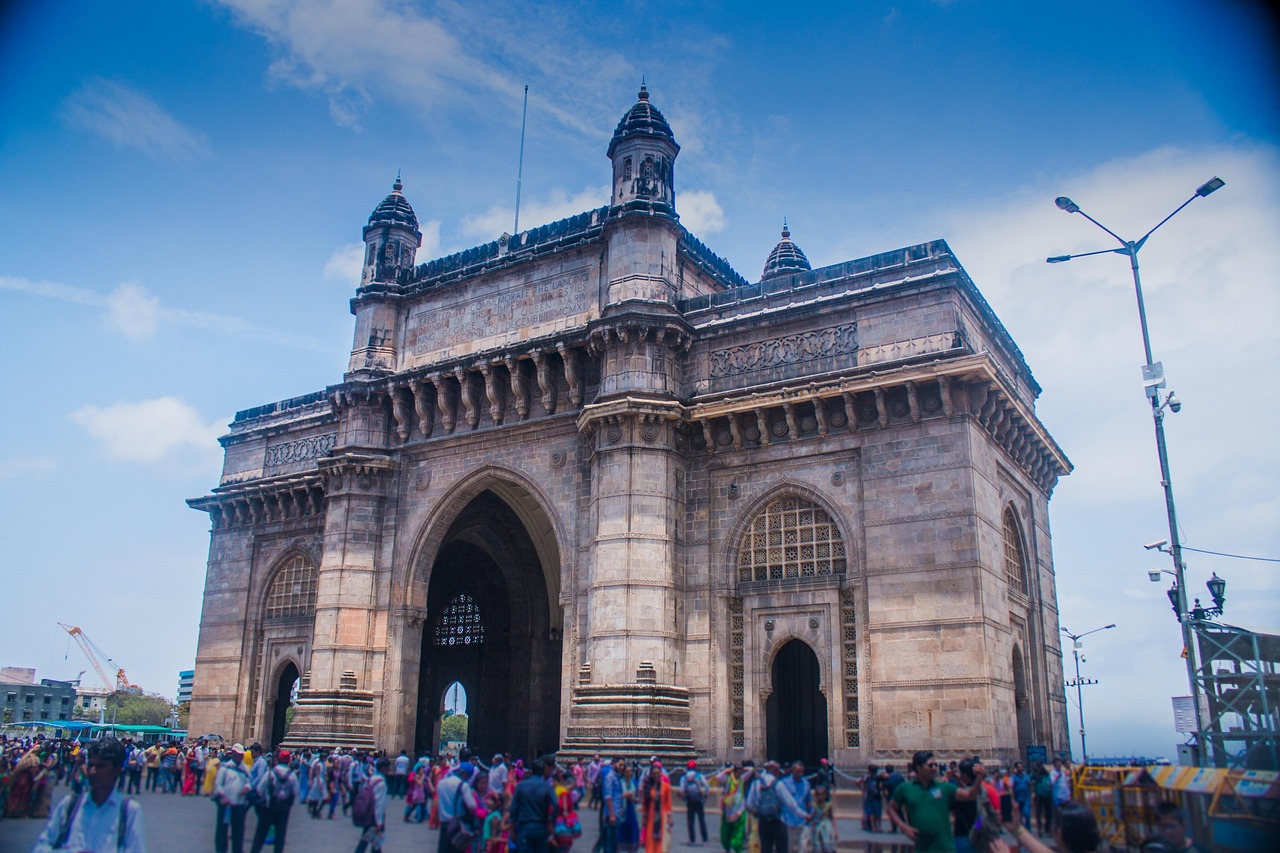 A large, ornate stone archway with intricate carvings and multiple arches, known as the Gateway of India, with many people walking in front of it.