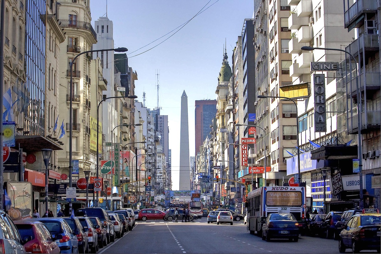 A bustling urban street in Buenos Aires with tall buildings, cars, and buses. The Obelisk of Buenos Aires is visible in the distance.