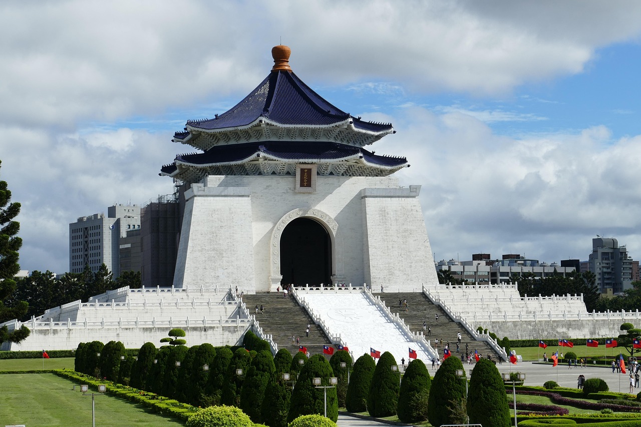 A grand white building with traditional Asian architecture, surrounded by green shrubbery and flags, set against a city skyline.