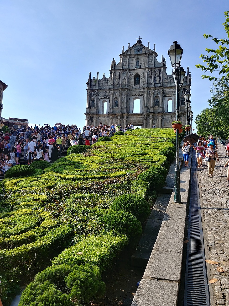 A large crowd of tourists gathered in front of the Ruins of St. Paul's in Macau, with well-maintained gardens and pathways leading up to the historic site.