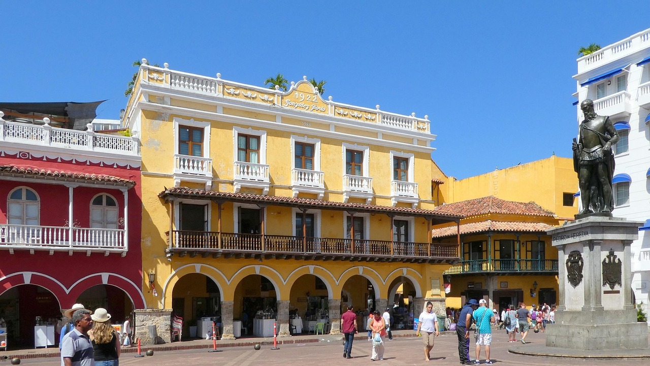 A bustling square in Cartagena, Colombia, with colorful colonial buildings, people walking, and a statue in the foreground.