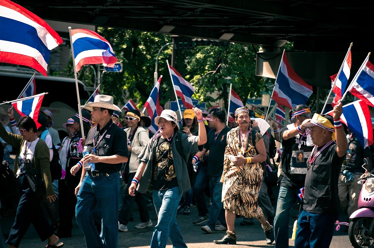A group of people marching in a parade or protest, holding Thai flags and wearing various attire.