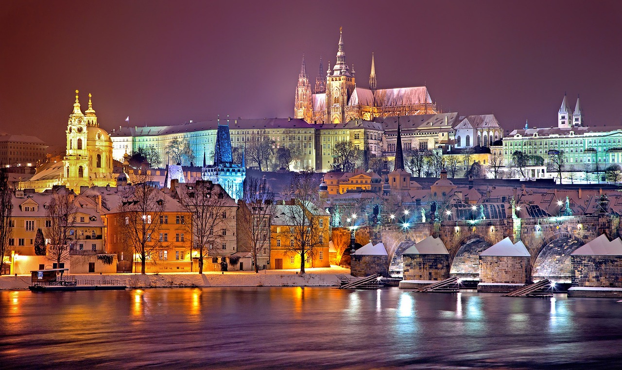 Night view of Prague Castle and the Charles Bridge in Prague, Czechia, with snow-covered buildings and a calm river in the foreground.