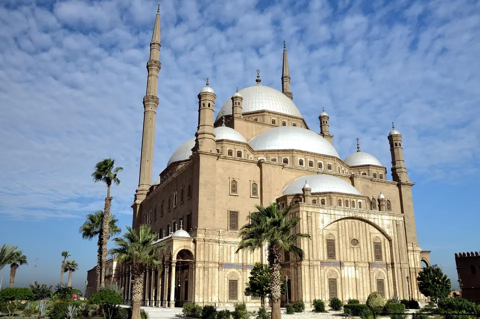 A large mosque with multiple domes and minarets under a clear blue sky with palm trees in the foreground.