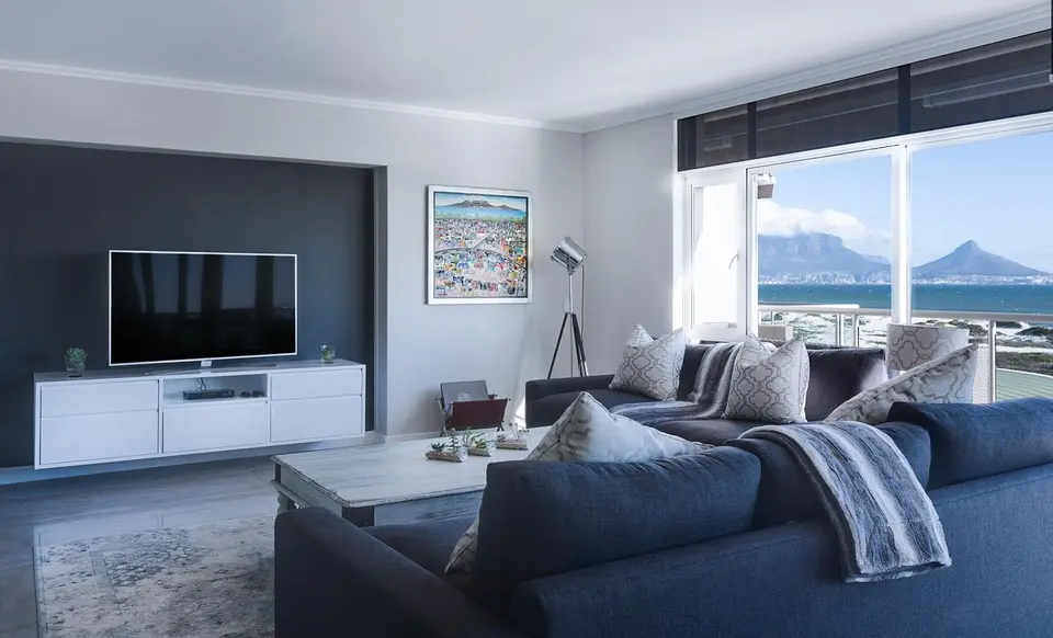 A modern living room with a sofa, coffee table, and a television. Large windows offer a view of a city and mountains.