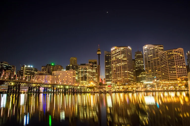 Nighttime cityscape of Sydney with illuminated skyscrapers and a bridge over the water.