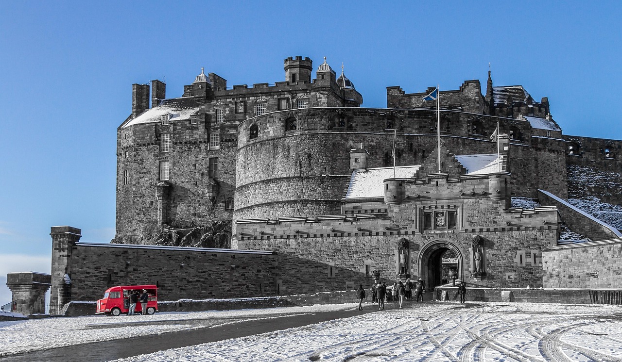 A large, historic castle with stone walls and towers, surrounded by snow. A red tourist vehicle is parked in front, and people are walking towards the entrance.