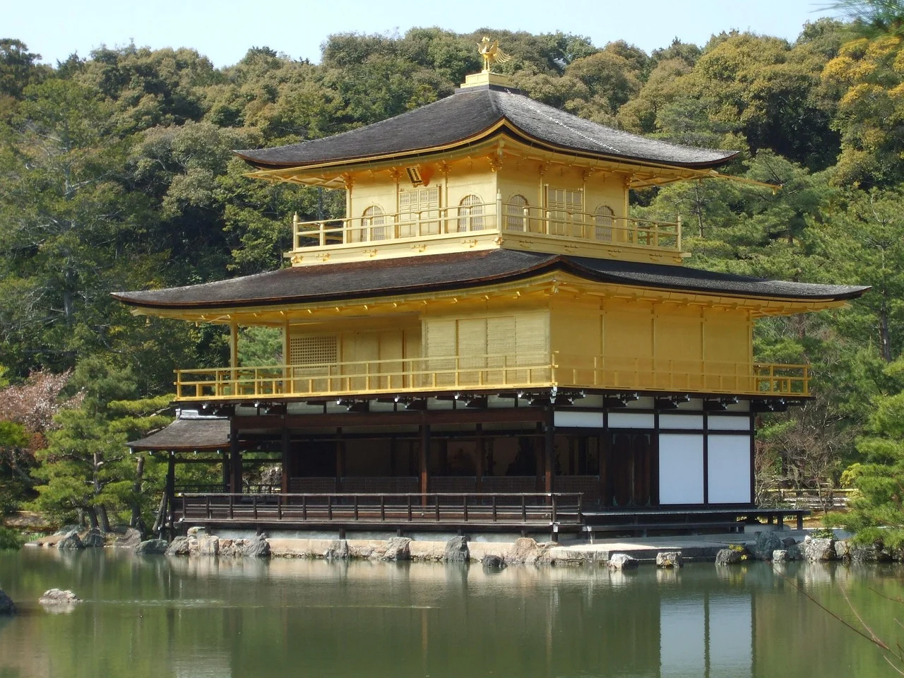 A golden pavilion with traditional Japanese architecture situated on a pond with lush green trees in the background.