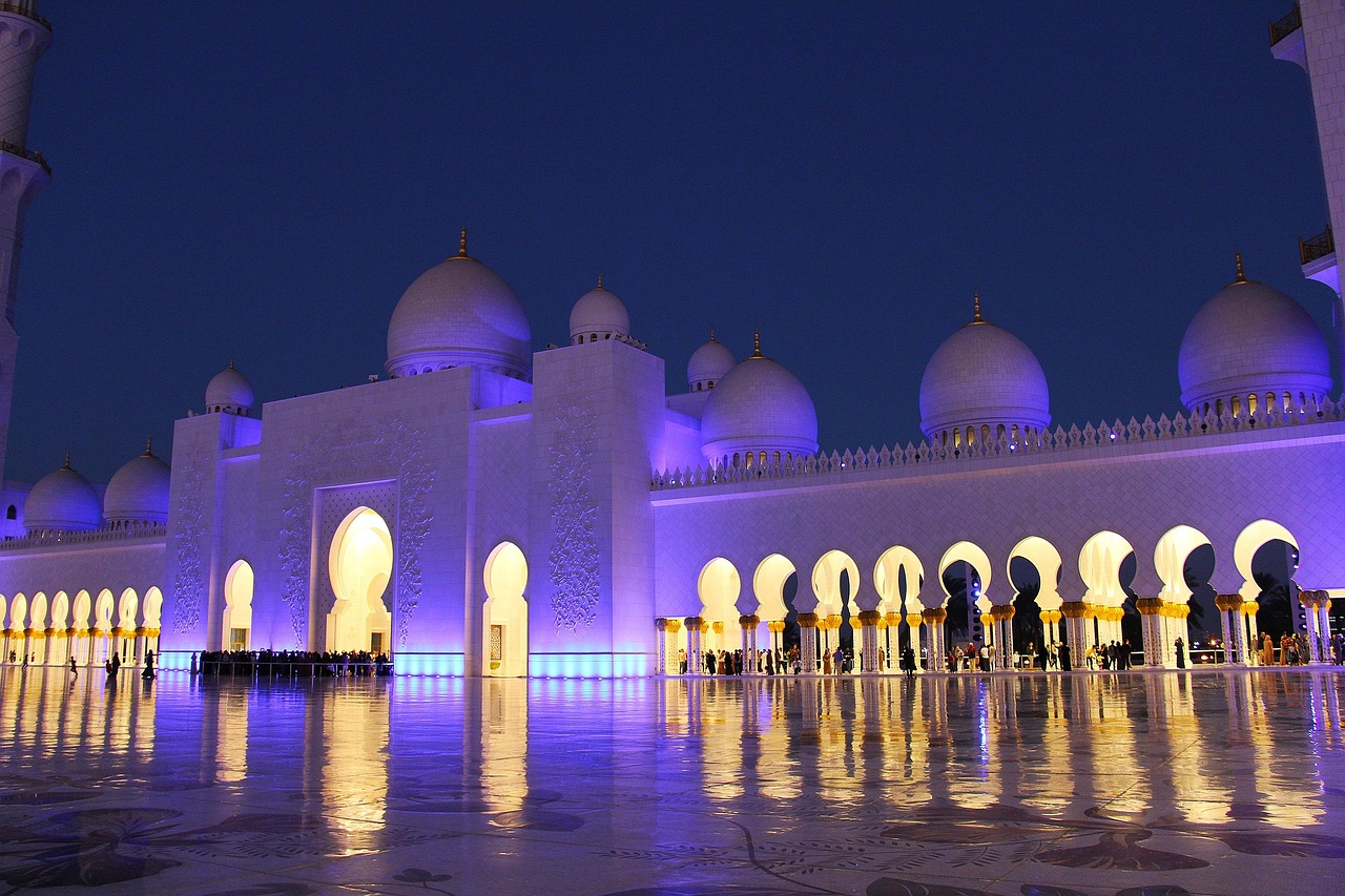 A night view of a large mosque with illuminated domes and arches, reflecting on a polished floor, with people gathered around.