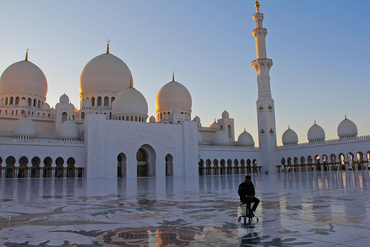 A person sitting on the ground in front of a large, white mosque with multiple domes and minarets during sunset.
