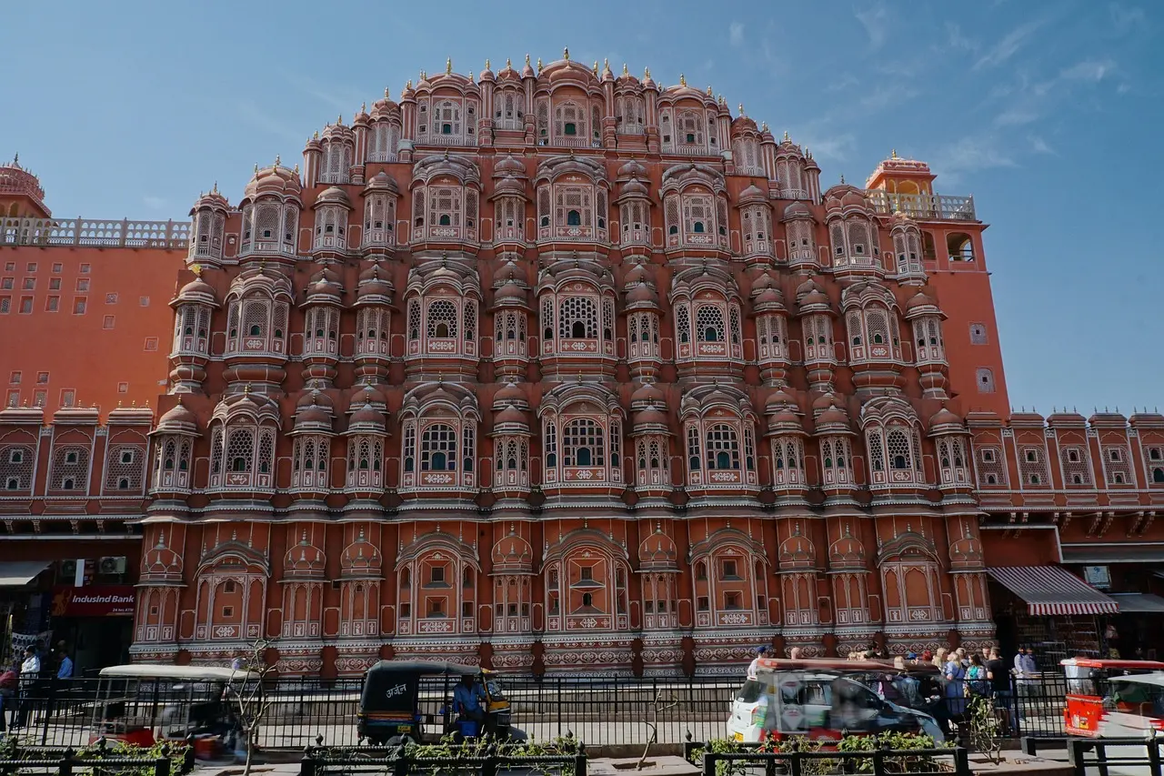 A large, ornate pink and white building with intricate designs, identified as the Hawa Mahal in Jaipur, India. The building has many windows and is bustling with people and vehicles in front.