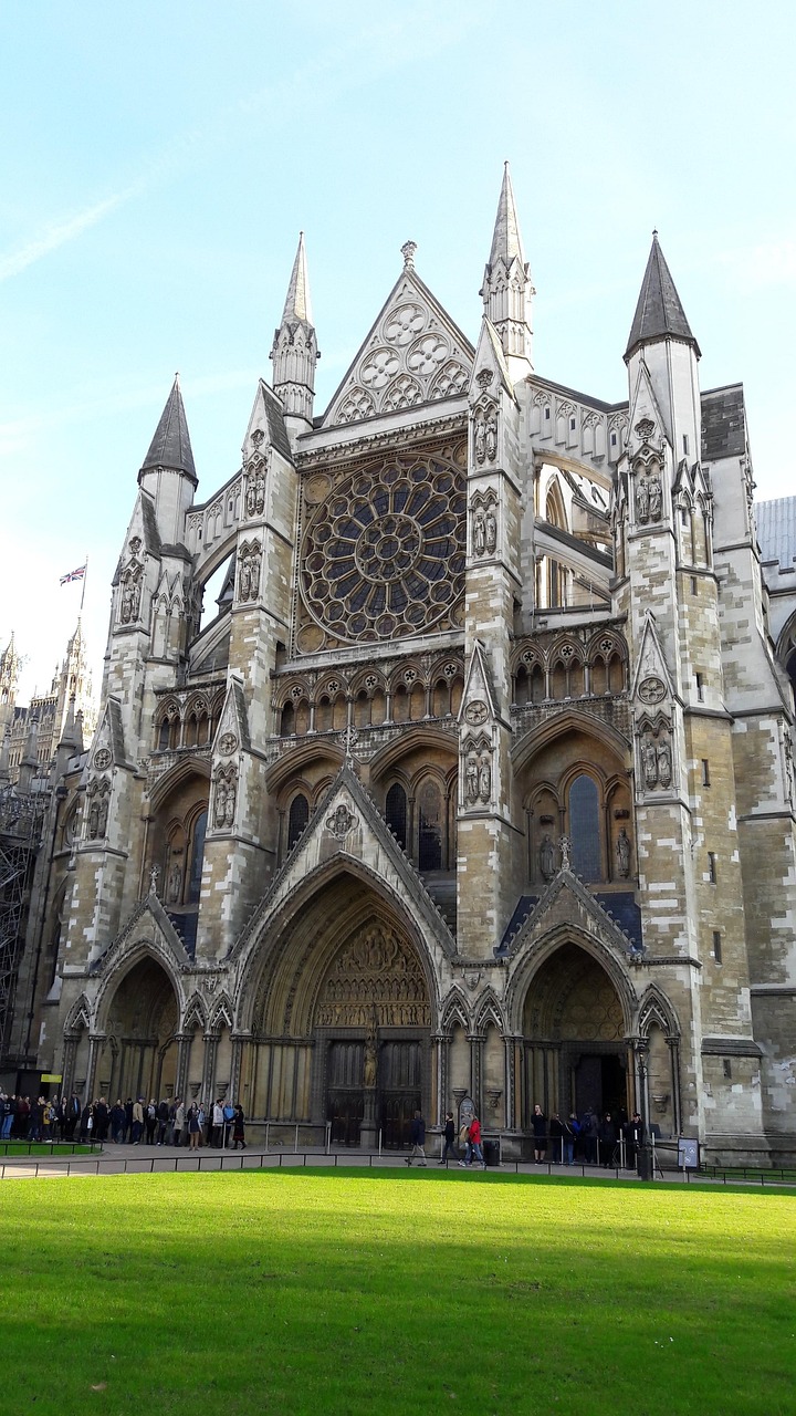 A grand Gothic cathedral with intricate details and large stained glass windows, surrounded by a green lawn and people gathered in front.