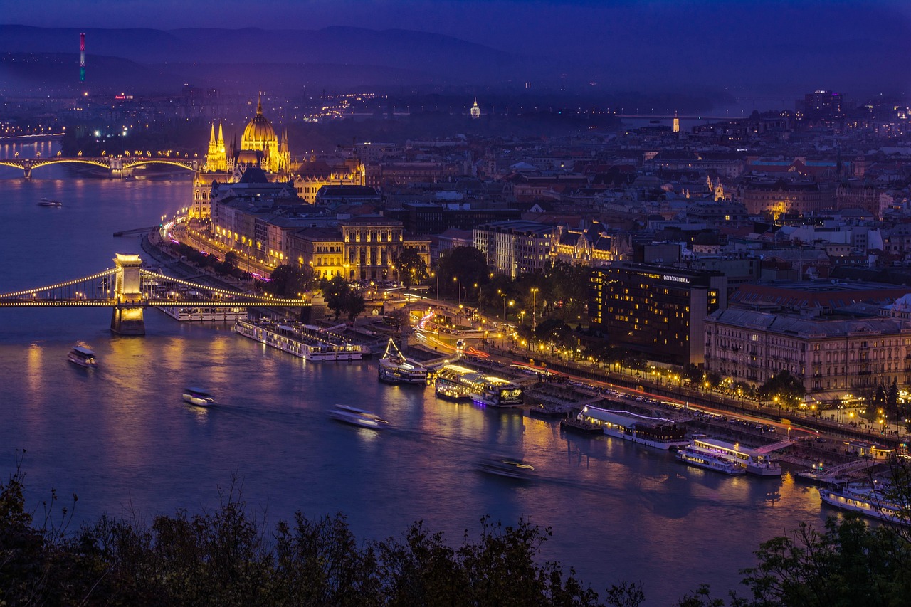 Night view of a city with a river, illuminated bridges, and boats, featuring distinctive architecture and a prominent dome-shaped building.