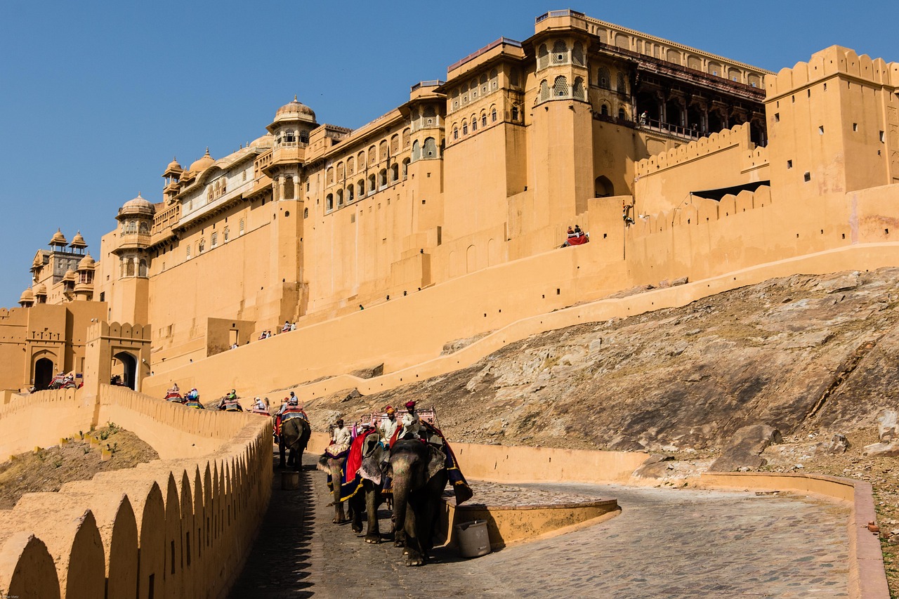 A large, historic fort with yellow sandstone walls, featuring elephants and tourists. The fort is situated on a hill with steps leading up to it.