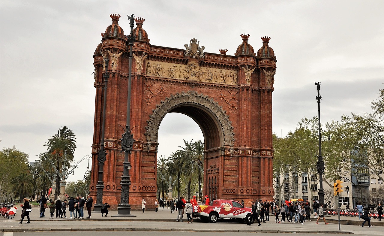 Large ornate archway with intricate designs and sculptures, surrounded by people and palm trees, with a red and white vehicle in front.