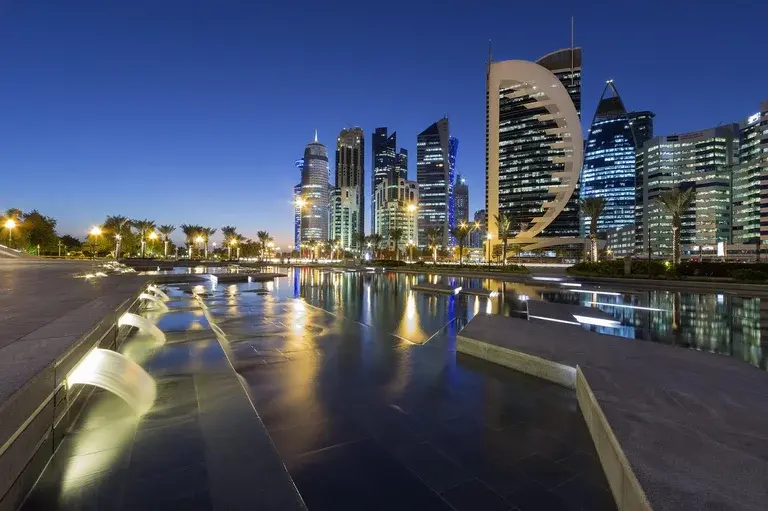Nighttime cityscape of Doha, Qatar, with illuminated skyscrapers and a reflective water feature in the foreground.