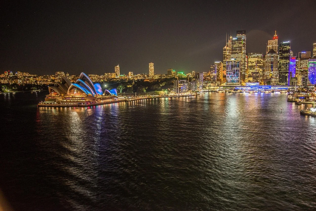 Night view of a city skyline with illuminated buildings and water in the foreground.