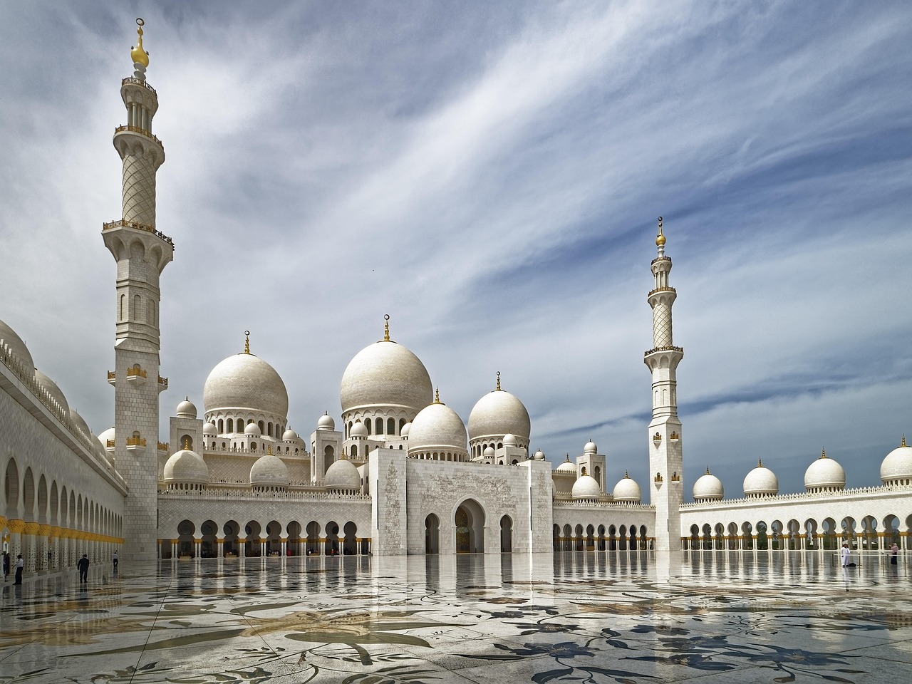 A grand mosque with large white domes and tall minarets, reflecting in a wet courtyard.
