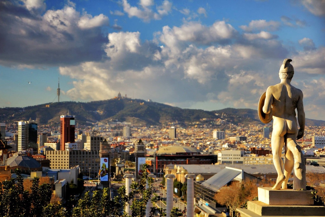A statue in the foreground with a cityscape and mountain in the background under a partly cloudy sky.