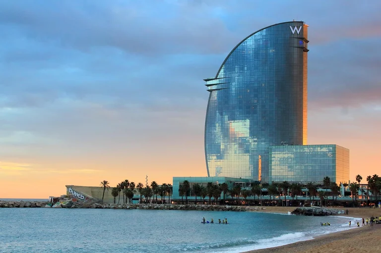 A modern hotel building with a distinctive sail-like shape at sunset, overlooking a beach with people swimming and walking along the shore.