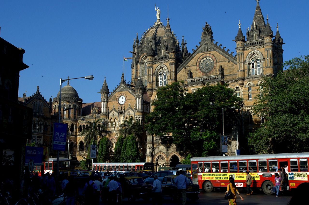 A bustling street scene in front of a large, ornate historical building with clock towers, featuring multiple buses and pedestrians.