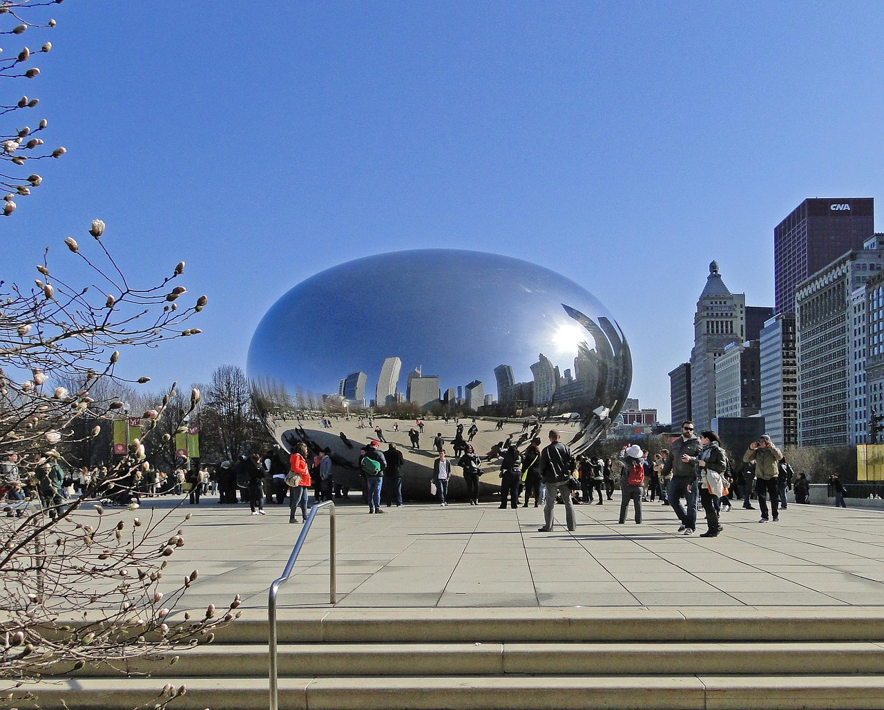 A large reflective bean-shaped sculpture in a public park with people walking around and tall buildings in the background.