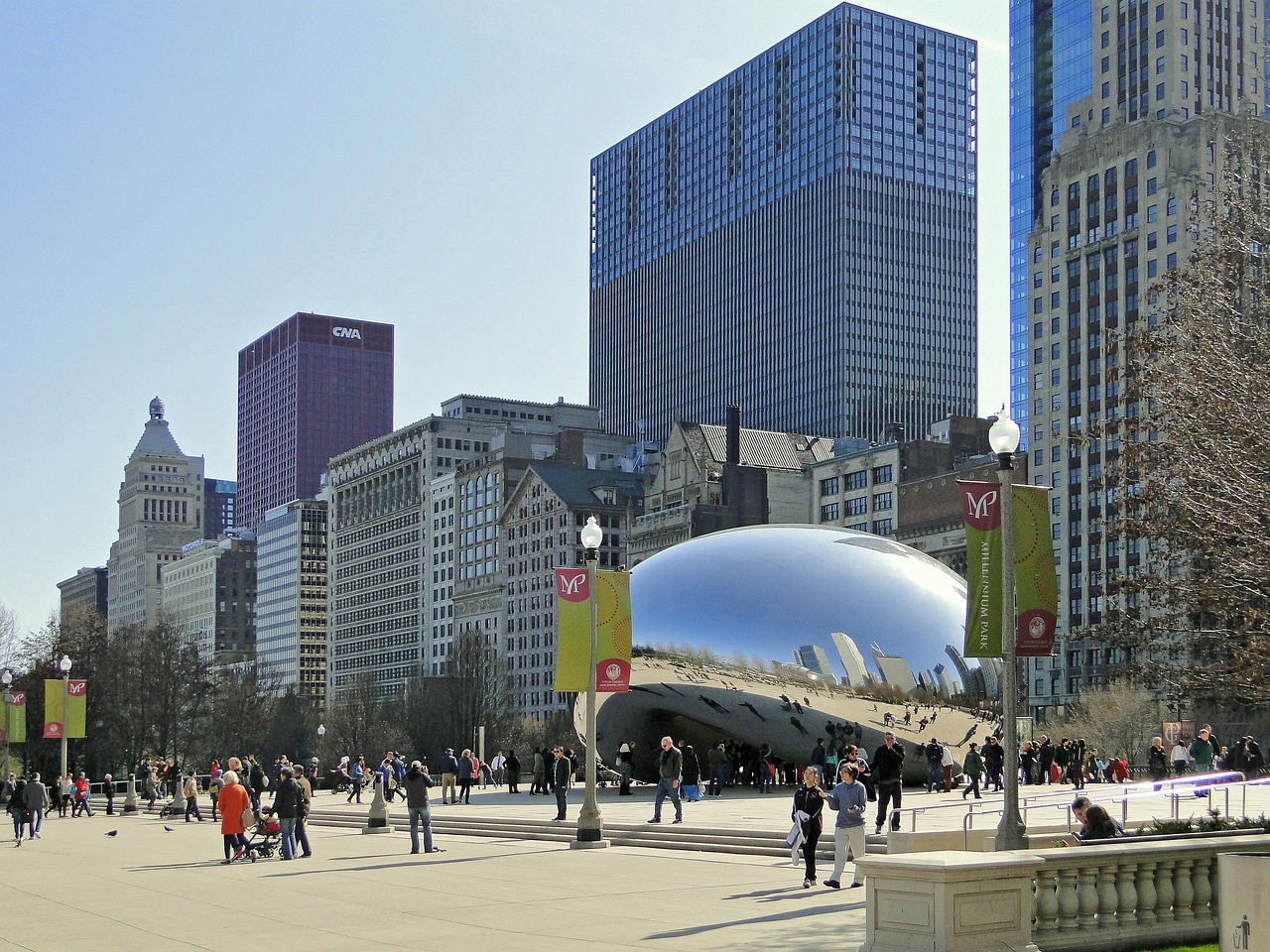 A large reflective bean-shaped sculpture in a public plaza surrounded by tall buildings and people walking around.