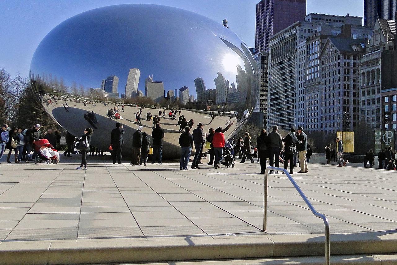 A large reflective bean-shaped sculpture in a city park with people walking around and tall buildings in the background.