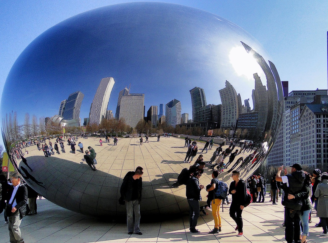 A large reflective bean-shaped sculpture in a public plaza with people around it and a city skyline in the background.