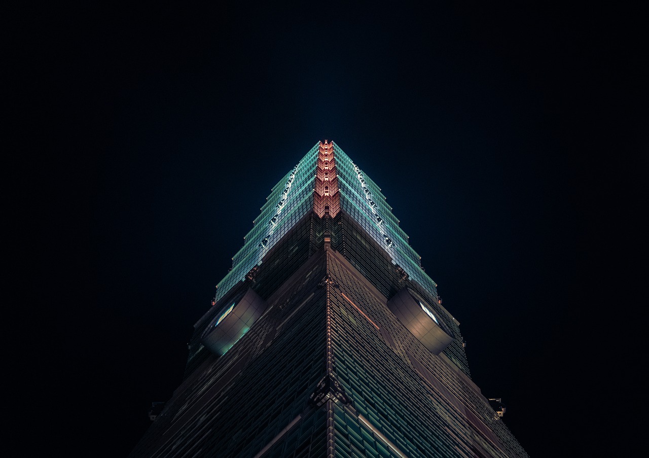 A night view of Taipei 101, a tall skyscraper with a distinctive tiered design, illuminated against a dark sky.